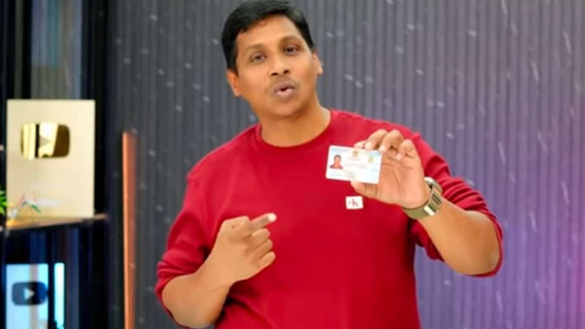 A man in a red sweatshirt holds up an identification card while speaking, with YouTube play buttons displayed in the background.
