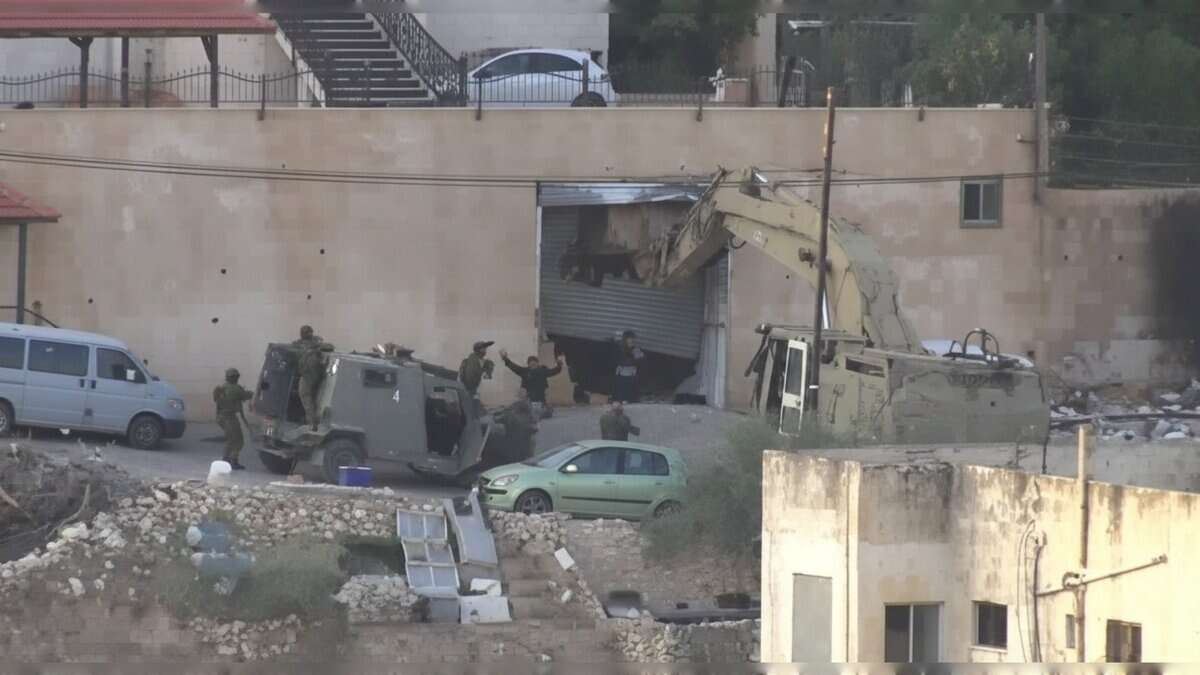 A group of Israeli soldiers stand outside a damaged building in Jenin as two Palestinian men emerge with their hands raised during a raid; a military bulldozer and armoured vehicles are positioned nearby.