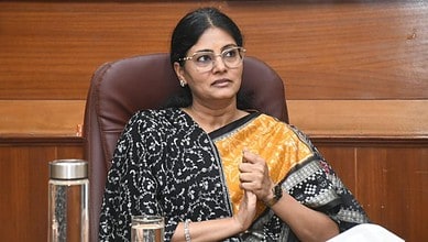 Anupriya Patel, Minister of State for Fertilisers, seated in an office chair wearing a black and orange patterned saree and glasses, with a water pitcher and glass on the desk