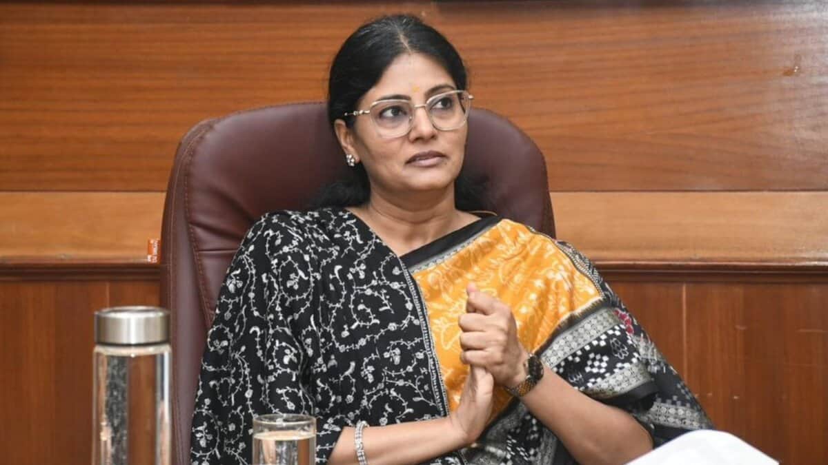 Anupriya Patel, Minister of State for Fertilisers, seated in an office chair wearing a black and orange patterned saree and glasses, with a water pitcher and glass on the desk