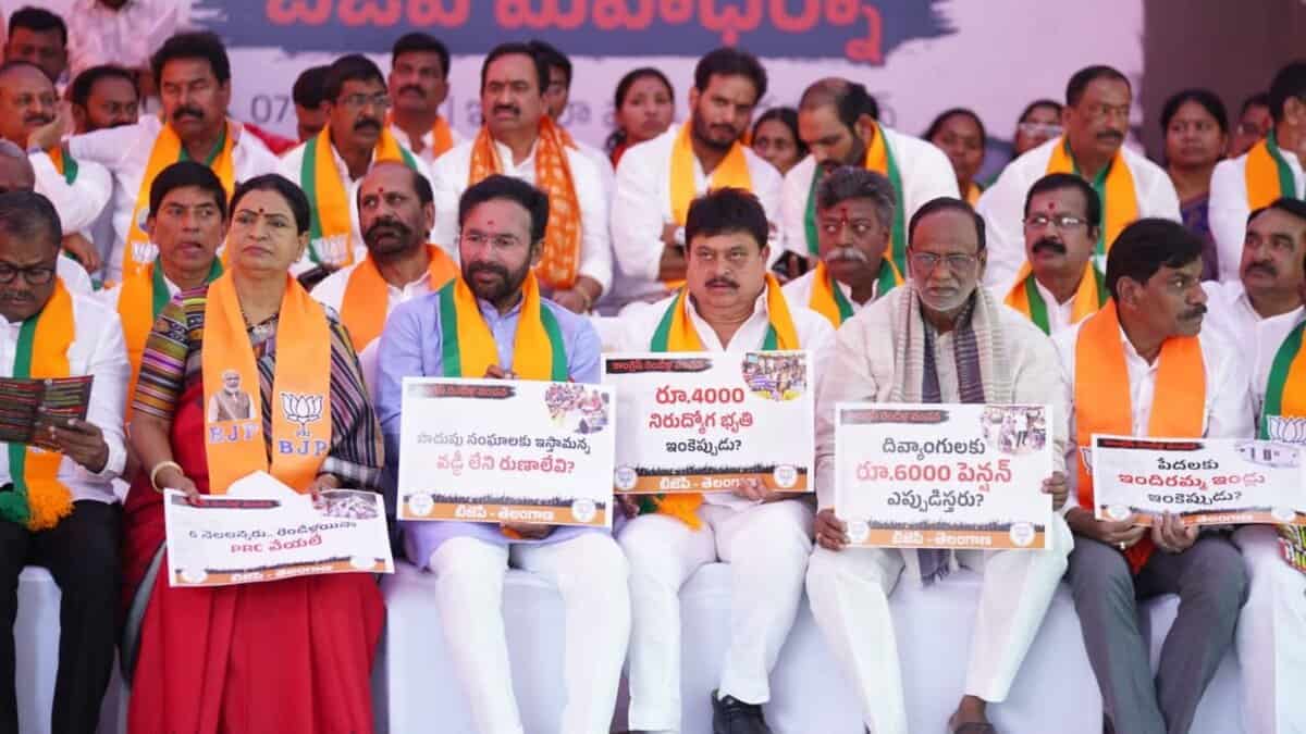 Political leaders wearing orange and green sashes sit on a stage holding signs with Telugu text and numerical figures during what appears to be a BJP party rally or demonstration.