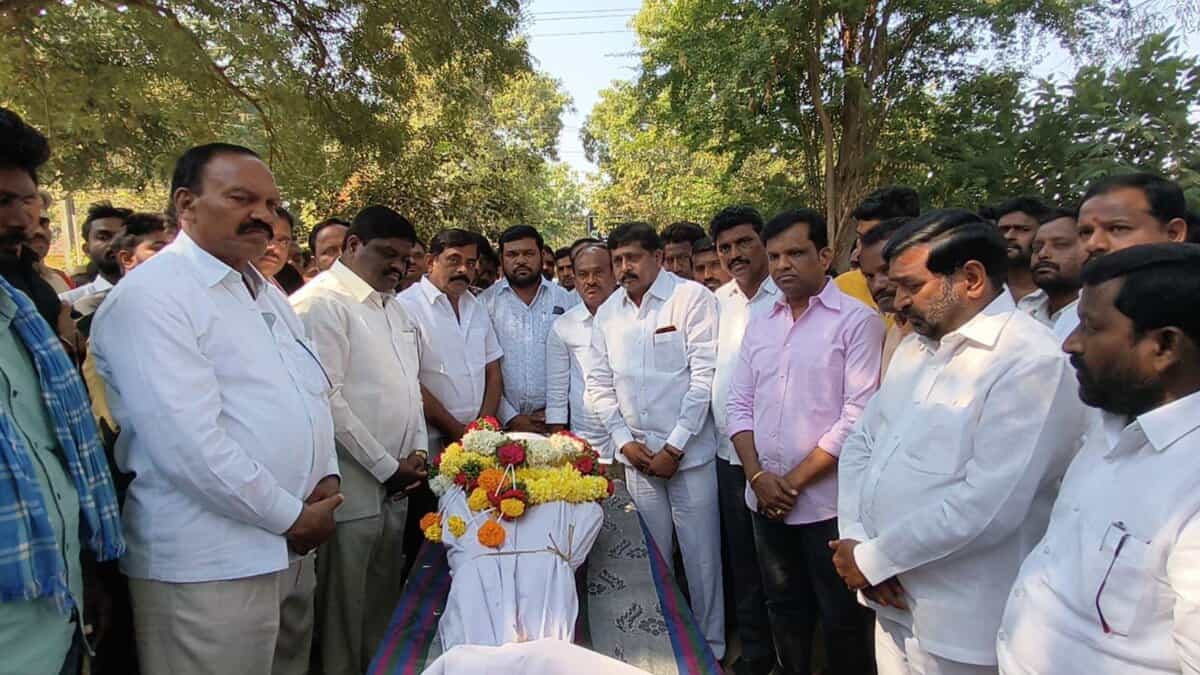 A group of men gather outdoors around a flower-draped casket during a funeral ceremony under trees.