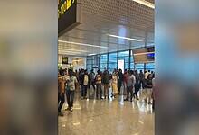 Crowded airport terminal with passengers gathered near gate 101, featuring modern architecture with large windows, textured ceiling panels, and polished floor tiles