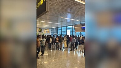 Crowded airport terminal with passengers gathered near gate 101, featuring modern architecture with large windows, textured ceiling panels, and polished floor tiles