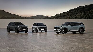 Three modern SUVs in dark gray, white, and light blue are displayed on a flat surface with rolling hills in the background at dusk.
