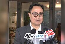 Kiren Rijiju in a dark suit and glasses speaking to media, with multiple microphones from ANI, IANS, and PTI news agencies positioned in front of him during a press conference