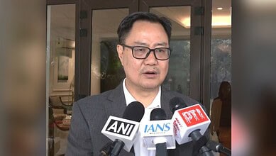 Kiren Rijiju in a dark suit and glasses speaking to media, with multiple microphones from ANI, IANS, and PTI news agencies positioned in front of him during a press conference