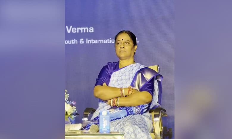 Woman in blue and white saree sitting on stage at a conference event, arms crossed, with microphone and backdrop visible behind her.