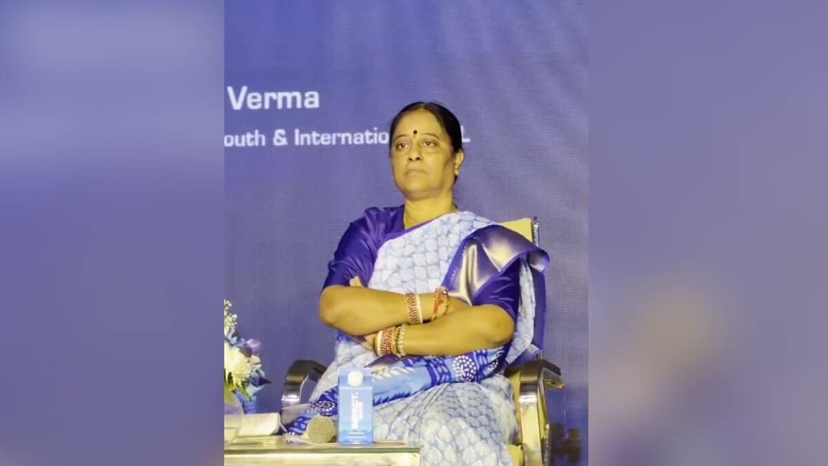 Woman in blue and white saree sitting on stage at a conference event, arms crossed, with microphone and backdrop visible behind her.