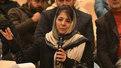 Mehbooba Mufti speaking into a microphone at a public event, wearing a beige and black embroidered headscarf, with attendees visible in the background