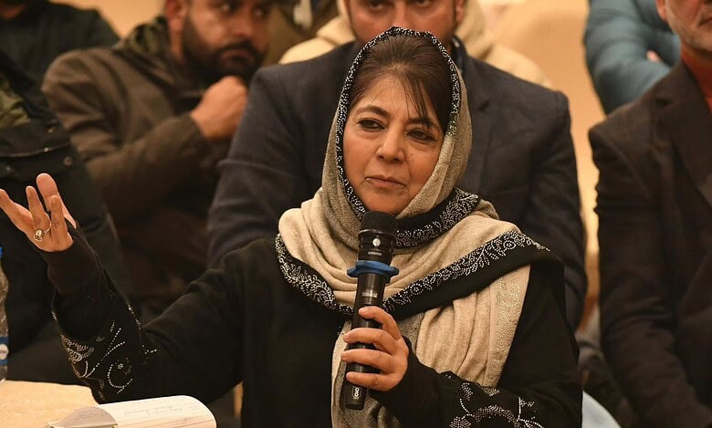 Mehbooba Mufti speaking into a microphone at a public event, wearing a beige and black embroidered headscarf, with attendees visible in the background