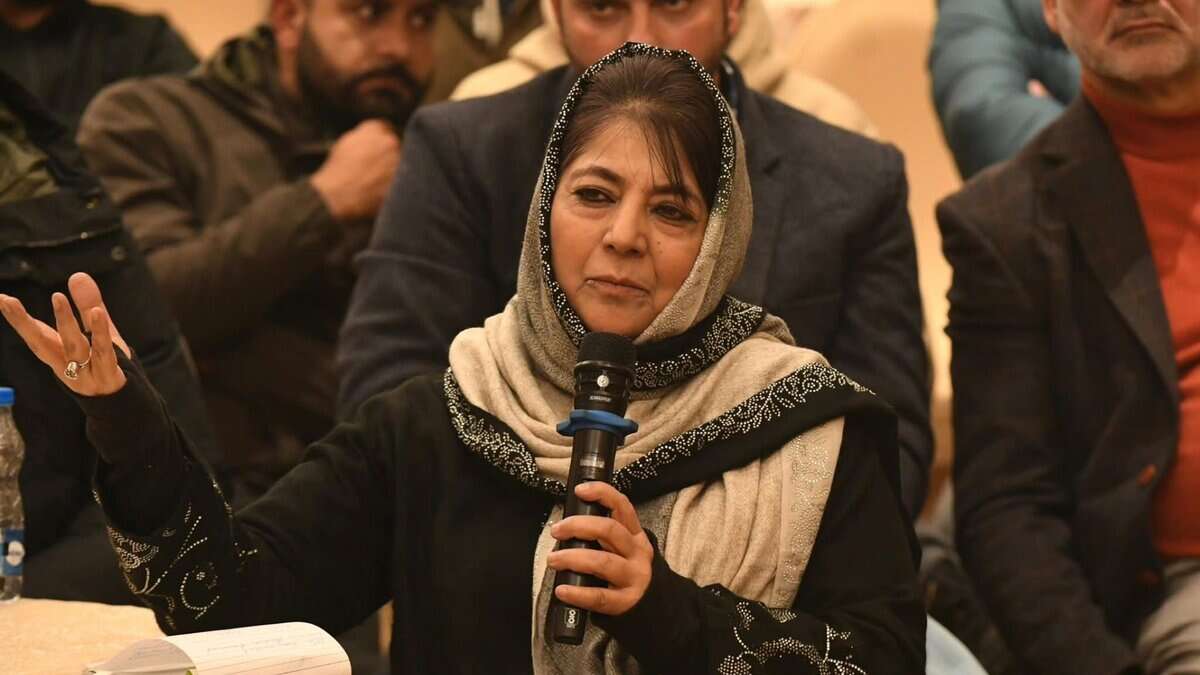 Mehbooba Mufti speaking into a microphone at a public event, wearing a beige and black embroidered headscarf, with attendees visible in the background
