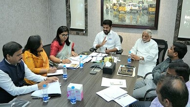 Government officials and team members seated around a conference table during a meeting, with documents, water bottles, and notebooks spread across the table, and a framed painting visible on the wall in the background