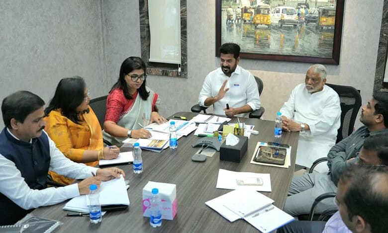Government officials and team members seated around a conference table during a meeting, with documents, water bottles, and notebooks spread across the table, and a framed painting visible on the wall in the background