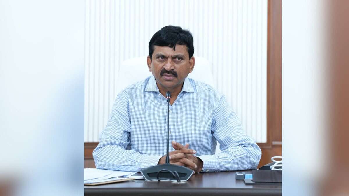 A man with a mustache wearing a light blue checkered shirt sits at a desk speaking into a microphone. He is gesturing with his hands while speaking. A conference phone and papers are visible on the desk in front of him, with a white curtained window in the background.