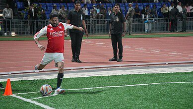 Revanth Reddy playing football on a field, wearing an Arsenal red and white jersey with 'Emirates Fly Better' sponsor, kicking a ball with officials and spectators visible in the background