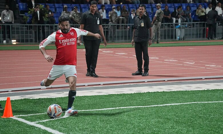 Revanth Reddy playing football on a field, wearing an Arsenal red and white jersey with 'Emirates Fly Better' sponsor, kicking a ball with officials and spectators visible in the background