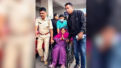 Woman in pink outfit sits in wheelchair with hands in namaste gesture, escorted by man in black jacket and police officer in khaki uniform through public building