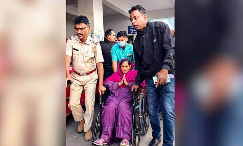 Woman in pink outfit sits in wheelchair with hands in namaste gesture, escorted by man in black jacket and police officer in khaki uniform through public building