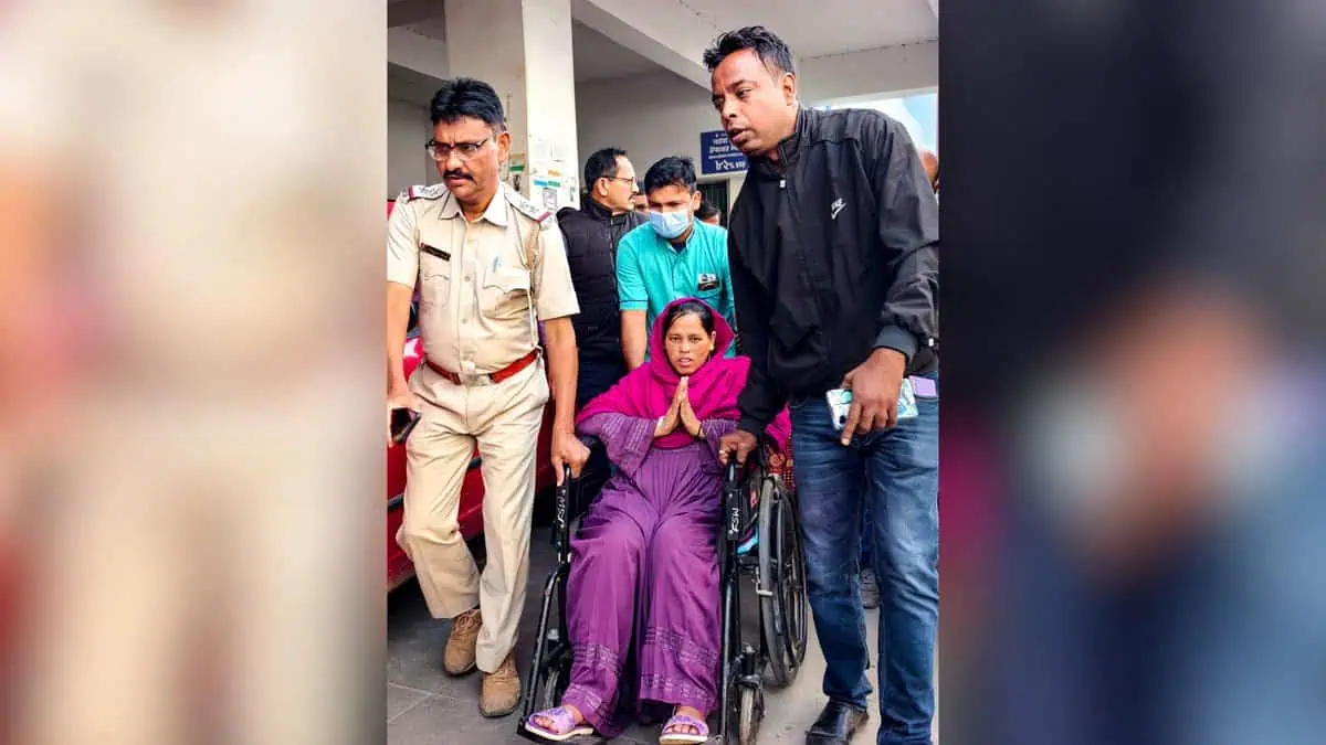 Woman in pink outfit sits in wheelchair with hands in namaste gesture, escorted by man in black jacket and police officer in khaki uniform through public building