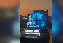 A large illuminated blue globe installation displays "Telangana Rising Global Summit" text against an urban skyline at dusk. The spherical structure features a world map design and is positioned on a waterfront platform with city buildings visible in the background.