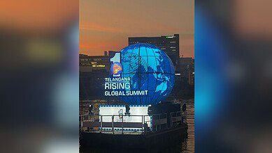 A large illuminated blue globe installation displays "Telangana Rising Global Summit" text against an urban skyline at dusk. The spherical structure features a world map design and is positioned on a waterfront platform with city buildings visible in the background.