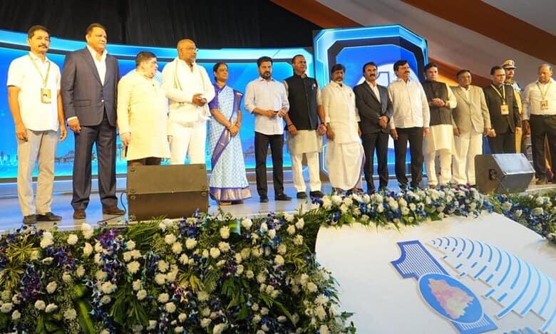 Large group of dignitaries standing on stage at a formal event with floral decorations and Telangana branding visible in the foreground.