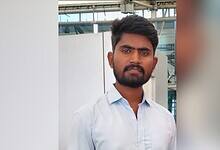 Young man with short dark hair and beard wearing a light blue shirt, photographed outdoors with industrial or commercial building structure visible in the background