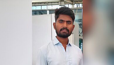 Young man with short dark hair and beard wearing a light blue shirt, photographed outdoors with industrial or commercial building structure visible in the background