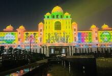 A grand government building illuminated at night with colorful projection mapping that displays vibrant yellow, green, pink, and blue patterns across its ornate facade. The architectural structure features multiple domes and is reflected in water below, with silhouettes of visitors visible in the foreground.