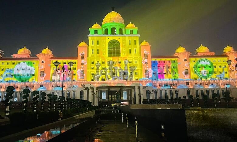 A grand government building illuminated at night with colorful projection mapping that displays vibrant yellow, green, pink, and blue patterns across its ornate facade. The architectural structure features multiple domes and is reflected in water below, with silhouettes of visitors visible in the foreground.