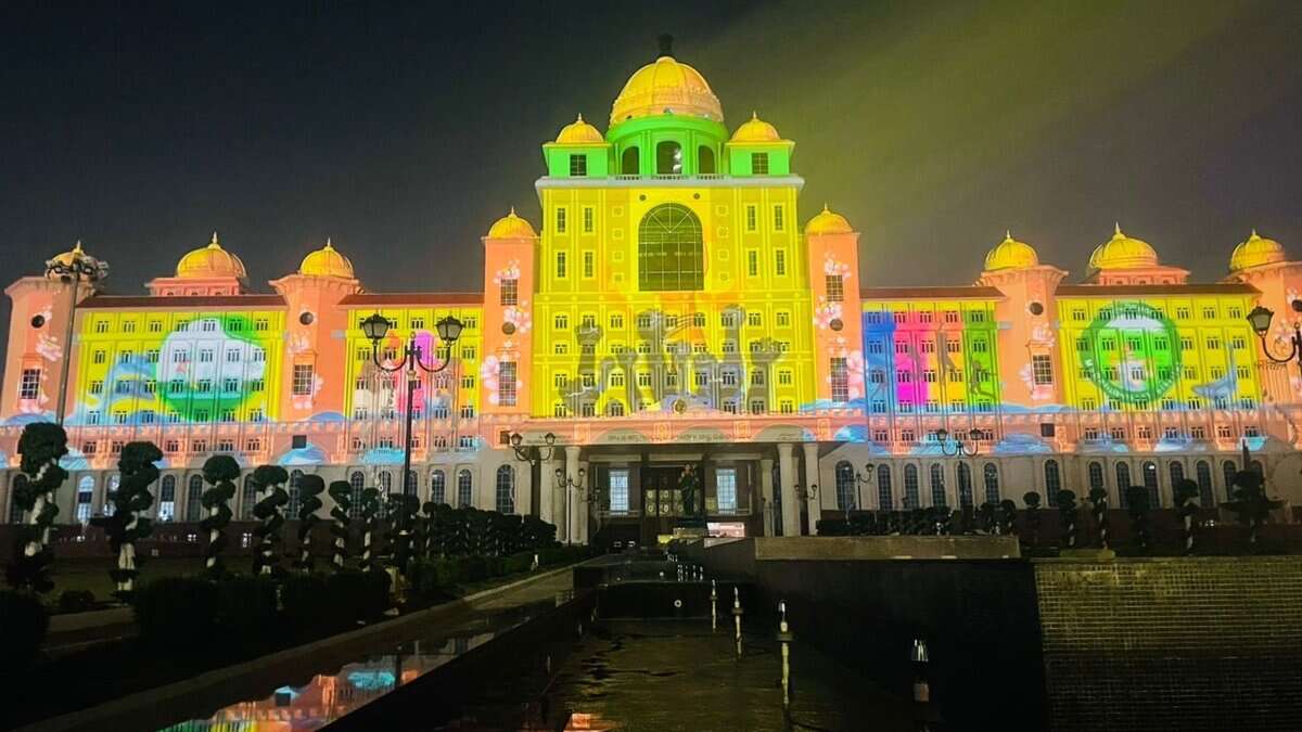 A grand government building illuminated at night with colorful projection mapping that displays vibrant yellow, green, pink, and blue patterns across its ornate facade. The architectural structure features multiple domes and is reflected in water below, with silhouettes of visitors visible in the foreground.