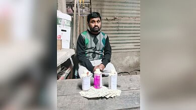 A man wearing a dark sweater with green and gray argyle pattern sits at a table with three bottles containing liquids (two clear, one pink) and Indian rupee notes arranged in front of him. The setting appears to be a small shop or workspace with corrugated metal walls and shelving visible in the background