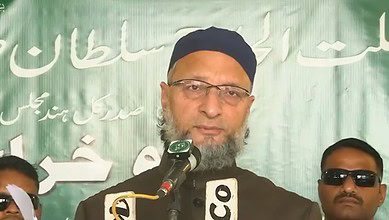 Man wearing black cap and glasses with a beard speaks at microphones during a press conference, flanked by two men in sunglasses. Green banner with Arabic and Urdu text visible in background.