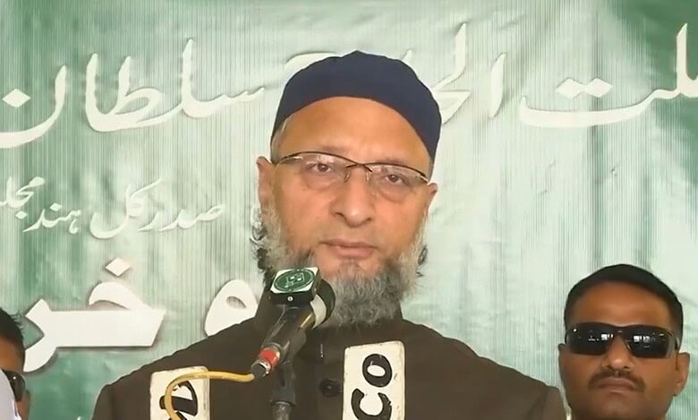 Man wearing black cap and glasses with a beard speaks at microphones during a press conference, flanked by two men in sunglasses. Green banner with Arabic and Urdu text visible in background.