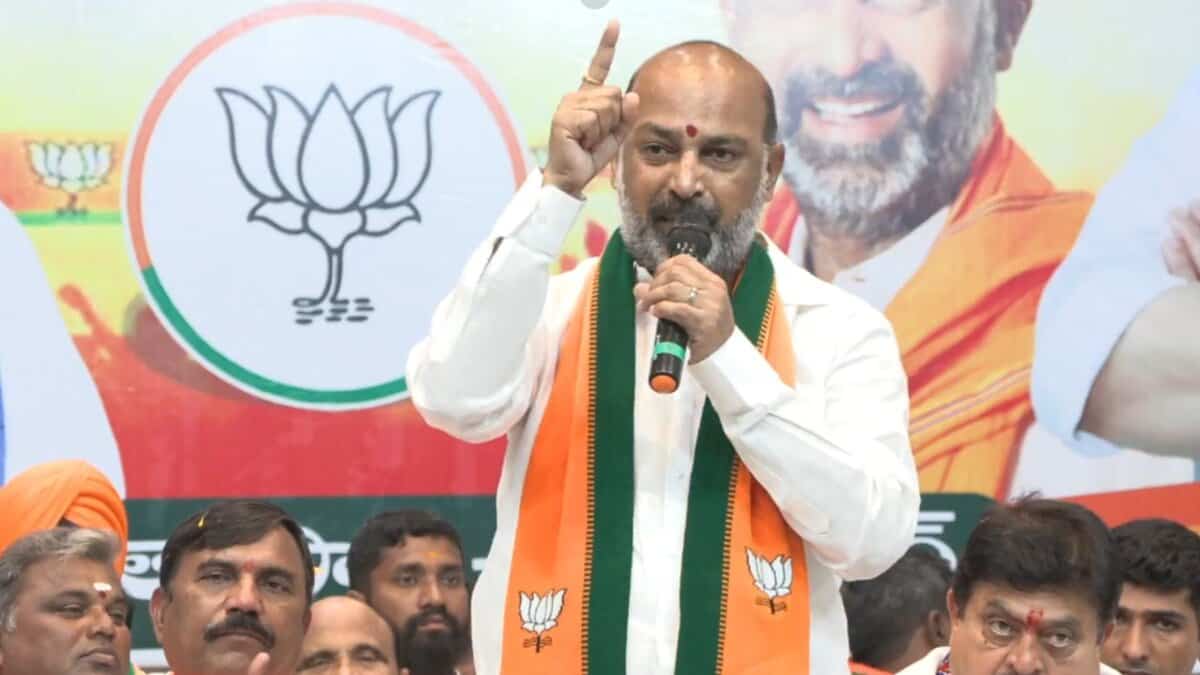 Bandi Sanjay in traditional white and saffron attire speaks passionately at a BJP event, gesturing with his finger raised while standing before the party's lotus symbol.