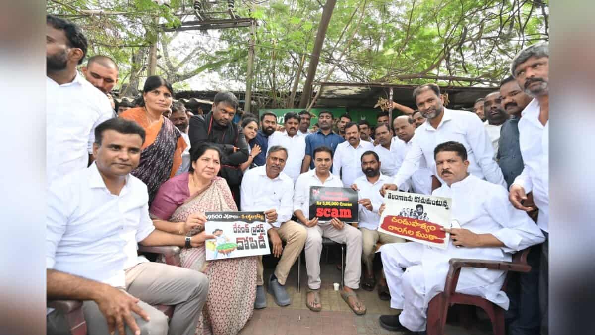 Political gathering showing BJP leaders and supporters sitting outdoors holding protest signs in Telugu about alleged scams. Group photo with men in white attire and a woman in saree, taken under trees with makeshift shelter in background.