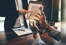 A person in handcuffs sits at a table during what appears to be a legal consultation, while someone in professional attire stands nearby holding a document. Law books and papers are visible on the wooden table surface.