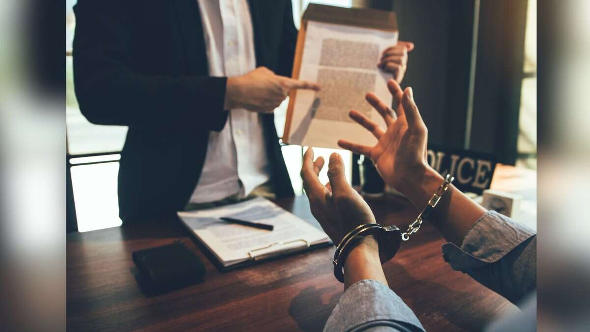 A person in handcuffs sits at a table during what appears to be a legal consultation, while someone in professional attire stands nearby holding a document. Law books and papers are visible on the wooden table surface.