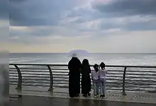 A woman and three children stand by the Jeddah waterfront under cloudy skies, looking out at the sea while holding an umbrella.