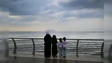 A woman and three children stand by the Jeddah waterfront under cloudy skies, looking out at the sea while holding an umbrella.