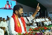 Political leader in white attire with red traditional shawl speaking at podium with raised finger gesture, addressing audience seated in background, with large screen display and floral arrangements visible