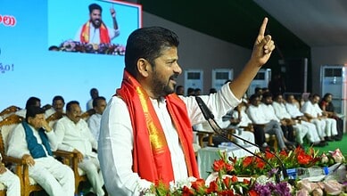 Political leader in white attire with red traditional shawl speaking at podium with raised finger gesture, addressing audience seated in background, with large screen display and floral arrangements visible