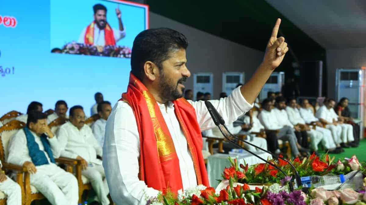 Political leader in white attire with red traditional shawl speaking at podium with raised finger gesture, addressing audience seated in background, with large screen display and floral arrangements visible