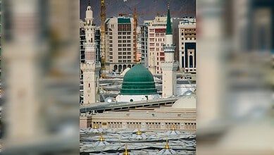 The green dome and minarets of the Prophet’s Mosque in Madinah, with surrounding buildings visible in the background.