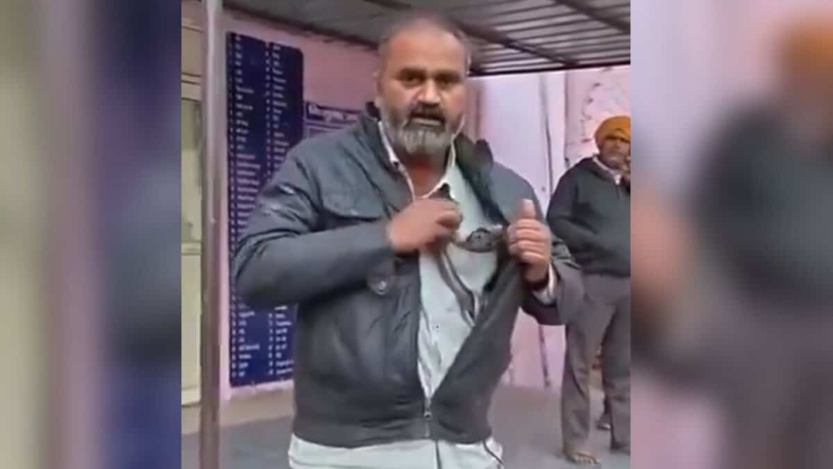 Image shows a man wearing a gray jacket and holding a snake stands in what appears to be a public space