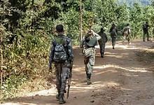 Image shows Armed soldiers in camouflage uniforms walking along a dirt road through a rural, tree-lined area.
