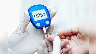 Image shows A healthcare worker in gloves performing a blood glucose test on a patient's finger with a blue glucometer