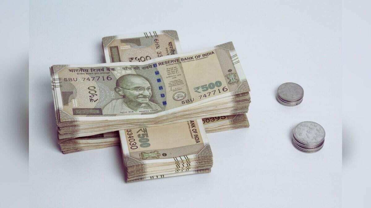 Image shows Stacks of Indian 500 rupee banknotes featuring Mahatma Gandhi alongside two piles of coins on a light background.