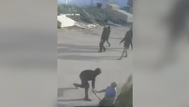 Image shows a masked man with a stick beating a man in a blue shirt